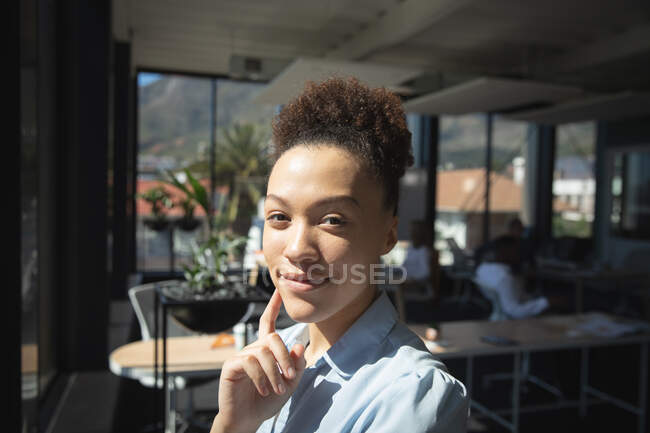 Retrato de una mujer de negocios de raza mixta trabajando en una oficina moderna, mirando a la cámara y sonriendo, con sus colegas trabajando en el fondo - foto de stock