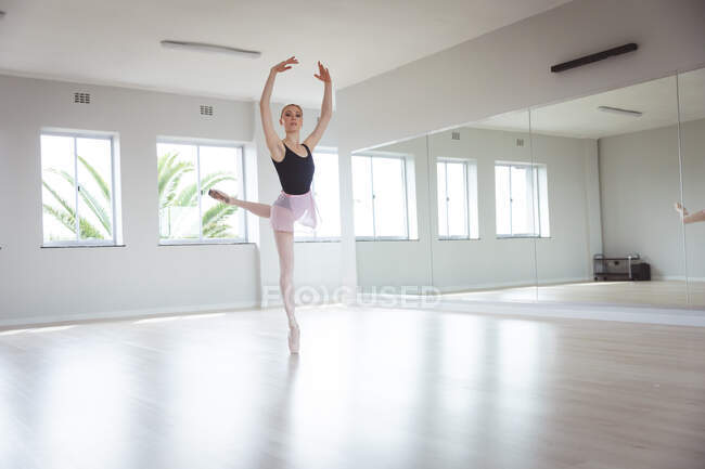 Кавказская привлекательная балетная танцовщица с рыжими волосами, танцующая в пуантах, стоящая на ногах во время балетной практики в яркой студии, сосредоточенная на упражнениях с руками над головой. — стоковое фото