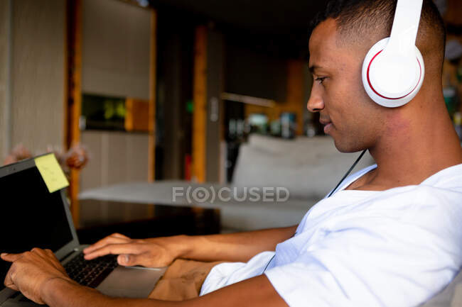 Vista lateral de perto de um homem afro-americano pendurado em sua sala de estar, sentado em um sofá, usando um computador portátil e usando fones de ouvido — Fotografia de Stock
