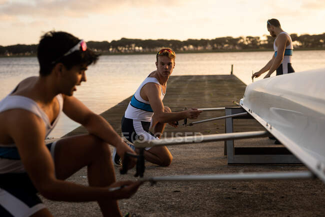 Side view of three Caucasian male rowers preparing a rowing boat before rowing, standing on a jetty on the river and talking — Stock Photo