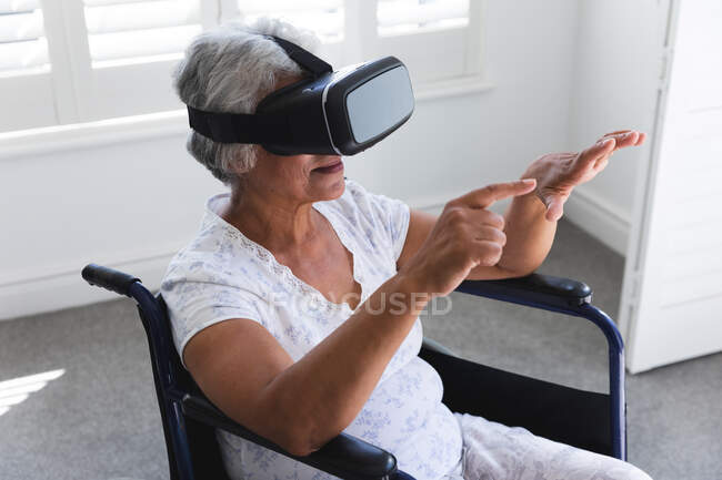 Eine ältere Afroamerikanerin im Ruhestand sitzt an einem sonnigen Tag im Rollstuhl vor einem Fenster und benutzt ein VR-Headset mit ausgestreckten Armen, um sich während der Coronavirus-Pandemie selbst zu isolieren 19 — Stockfoto