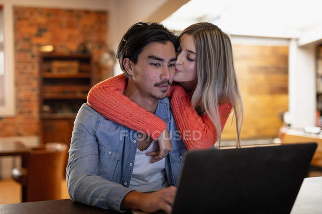 Передній погляд на молоду кавказьку жінку і молодого чоловіка змішаної раси, що відпочиває вдома, сидить у вітальні, посміхається і обіймає, використовуючи ноутбук, жінка цілує чоловіка в щоку.. — стокове фото