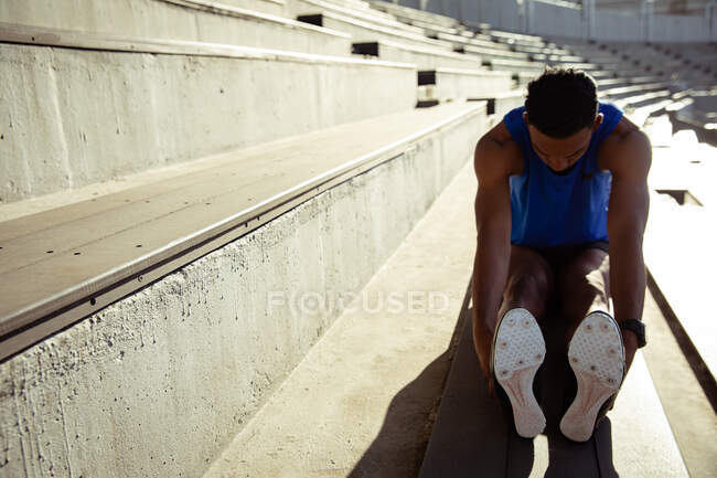Vista frontal de un atleta masculino de raza mixta practicando en un estadio deportivo, sentado en las gradas y estirándose. Entrenamiento deportivo de pista y campo en el estadio. - foto de stock