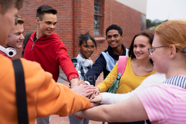 Front view of a multi-ethnic group of teenage male and female high school students with schoolbags hanging out, standing and hand stacking in their school grounds — Stock Photo