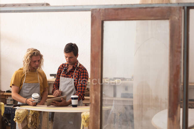 Two Caucasian male surfboard makers standing in their studio, preparing for work, discussing the project, using a tablet computer and holding cups of takeaway coffee. — Stock Photo