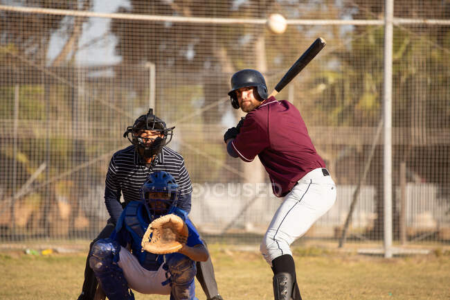 Vista frontal de un jugador de béisbol masculino caucásico durante un juego de béisbol en un día soleado, preparándose para golpear una pelota con un bate de béisbol, un receptor y otro jugador están sentados detrás de un bateador - foto de stock