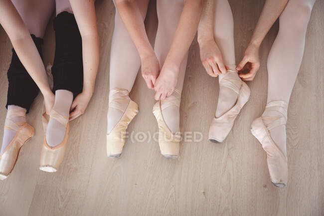 Section basse d'un groupe de danseuses de ballet attachant leurs chaussures de ballet dans un studio de ballet lumineux, préparant un cours de ballet, assises sur le sol. — Photo de stock