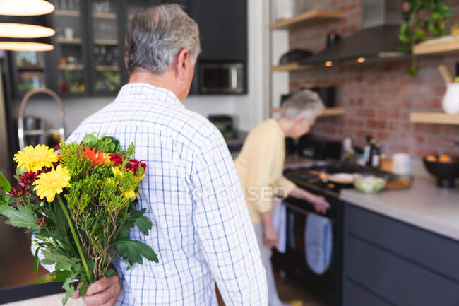 Visão traseira do casal caucasiano sênior desfrutando de sua aposentadoria, de pé em sua cozinha em um dia ensolarado, a mulher cozinhando e o homem segurando um buquê de flores atrás de suas costas — Fotografia de Stock