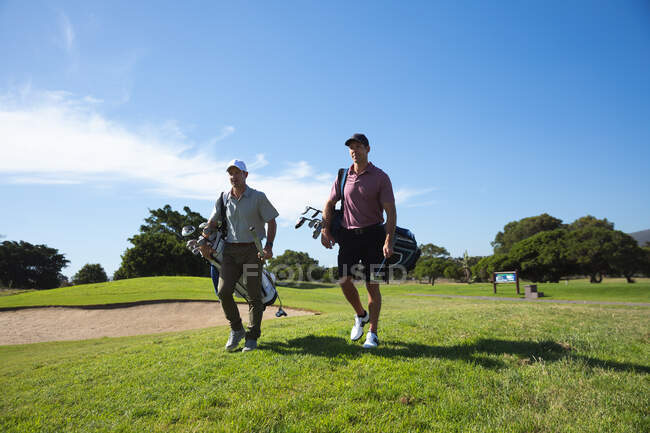 Frontansicht zweier kaukasischer Männer auf einem Golfplatz an einem sonnigen Tag mit blauem Himmel, beim Wandern, Golftaschen tragend — Stockfoto