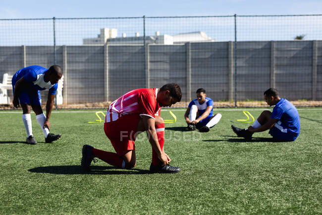 Multi etnia maschile cinque giocatori di calcio di lato che indossano un allenamento di striscia di squadra in un campo sportivo al sole, riscaldandosi legando i lacci delle scarpe. — Foto stock