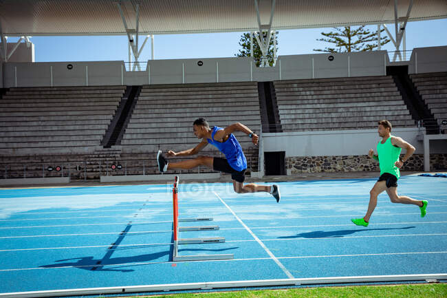 Seitenansicht eines kaukasischen und eines gemischten Rennens männliche Athleten, die in einem Sportstadion üben, sich gegenseitig auf der Laufbahn, beim Hürdenlauf — Stockfoto