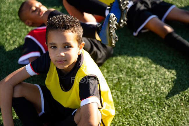 High angle portrait of a young mixed race boy soccer player sitting on a playing field in the sun and looking up to camera during a soccer training session, with his teammates training in the background — Stock Photo