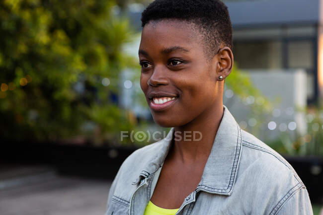 Vista lateral de perto de uma mulher afro-americana pendurada em um terraço em um dia ensolarado, olhando para longe da câmera e sorrindo — Fotografia de Stock
