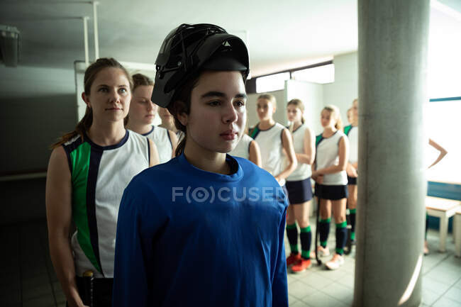 Side view of a Caucasian female field hockey players, preparing before a game, standing in a changing room, wearing a hockey helmet, with her teammates standing in a row behind her — Stock Photo