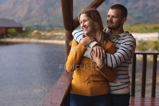 Seitenansicht eines kaukasischen Paares, das eine gute Zeit auf einer Reise in die Berge hat, auf einem Balkon in einer Hütte steht, sich umarmt, die Aussicht genießt — Stockfoto