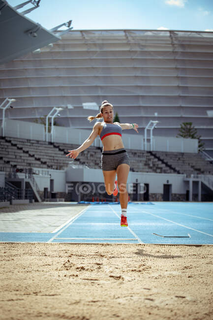Vista frontale di un'atleta caucasica che pratica in uno stadio sportivo, facendo un salto in lungo. — Foto stock