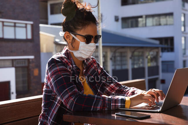 Seitenansicht einer Mischlingsfrau mit langen dunklen Haaren, die tagsüber an einem Tisch in einem Café sitzt, Sonnenbrille und Mundschutz gegen Luftverschmutzung und Coronavirus trägt und an einem Laptop mit Gebäuden im Hintergrund arbeitet. — Stockfoto