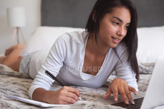 Mixed race woman spending time at home self isolating and social distancing in quarantine lockdown during coronavirus covid 19 epidemic, lying on bed using laptop making notes in bedroom. — Stock Photo