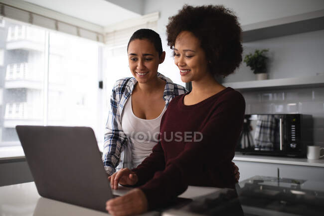 Вид спереду крупним планом змішаної раси жіноча пара розслабляється вдома, стоячи на кухні, використовуючи ноутбук комп'ютер разом і посміхаючись — стокове фото