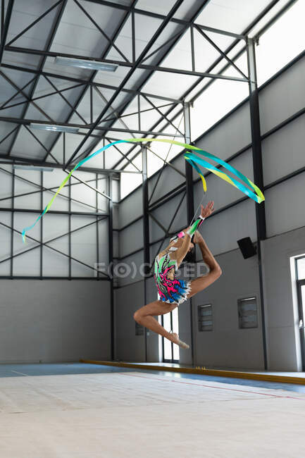 Vista lateral de la gimnasta femenina adolescente de raza mixta que actúa en el gimnasio, haciendo ejercicio con cinta, saltando, usando un maillot multicolor - foto de stock