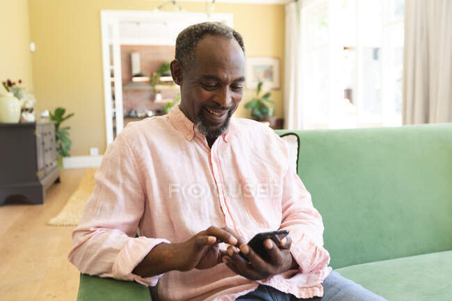Heureux beau retraité afro-américain à la maison assis sur le canapé dans son salon, à l'aide d'un smartphone et souriant, auto-isolant pendant la pandémie de coronavirus covid19 — Photo de stock