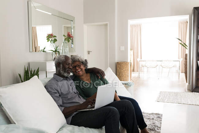 Старшая афроамериканская пара проводит время дома вместе, социальное дистанцирование и самоизоляция в карантинной изоляции во время эпидемии коронавируса, сидя на диване, обнимаясь, используя ноутбук — стоковое фото