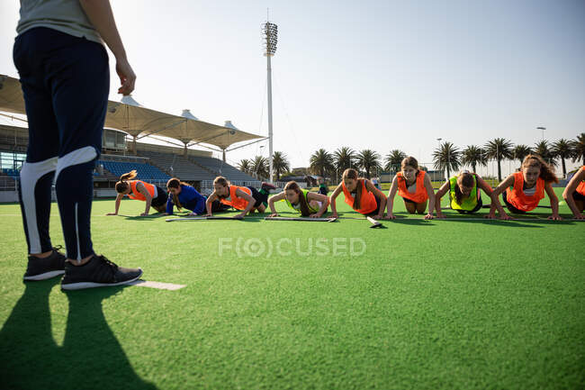 Front view of a group of female Caucasian field hockey players, training before a game, working out on a field hockey pitch, doing push ups, with their coach in the foreground, on a sunny day — Stock Photo
