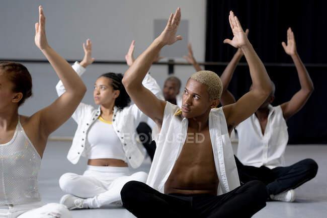 Vista frontale da vicino di un gruppo multietnico di ballerine moderne e maschili in forma con indosso abiti bianchi che praticano una routine di danza durante una lezione di danza in uno studio luminoso, sedute sul pavimento con le mani in alto. — Foto stock