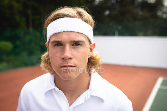 Retrato de un hombre caucásico con ropa blanca de tenis que pasa tiempo en una cancha jugando al tenis en un día soleado, mirando a la cámara - foto de stock