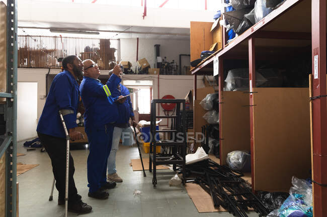 Three of Caucasian and African American male workers in a storage warehouse at a factory making wheelchairs, standing and talking, checking parts on shelves, two of them are disabled and using crutches — Stock Photo