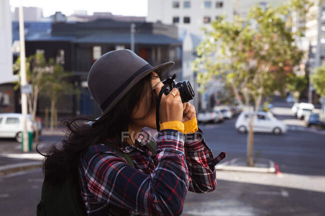 Vista lateral de una mujer feliz de raza mixta con el pelo largo y oscuro por las calles de la ciudad durante el día, llevando una mochila, usando un sombrero y una camisa revisada mirando a través de prismáticos con edificios en el fondo. - foto de stock