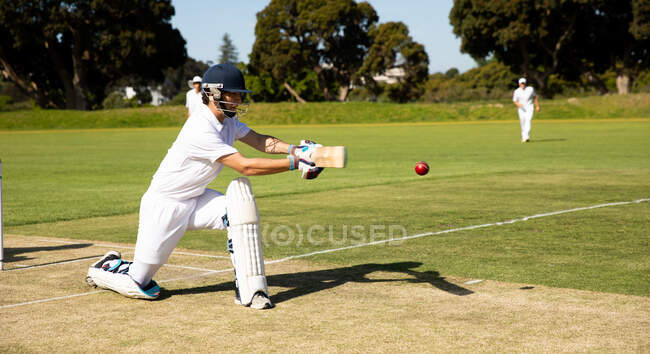 Vista lateral de um adolescente caucasiano jogador de críquete ajoelhado no campo usando capacete e luvas, segurando um taco de críquete, batendo a bola durante uma partida de críquete, com outros jogadores correndo em segundo plano. — Fotografia de Stock