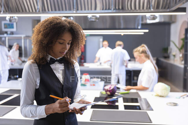 Mixed race waitress writing on a notebook, with chefs cooking and cutting vegetables in the background. Cookery class at a restaurant kitchen. — Stock Photo