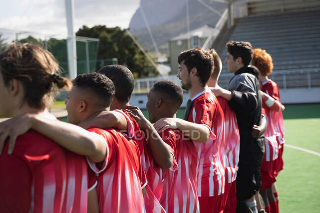 Side view of a multi-ethnic group of male field hockey players and their Caucasian male coach, preparing before a game, standing in a row on the pitch with their arms around each others shoulders on a sunny day — Stock Photo