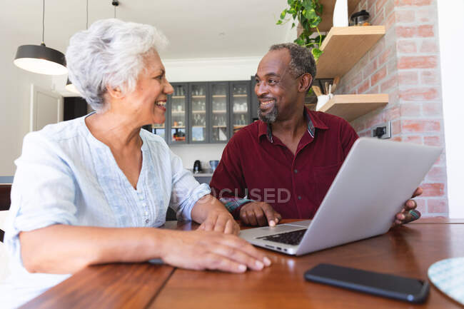Primer plano de una feliz pareja de ancianos afroamericanos jubilados sentados en una mesa en su comedor, usando una computadora portátil juntos y sonriéndose mutuamente, en casa juntos aislándose durante la pandemia de coronavirus covid19 - foto de stock