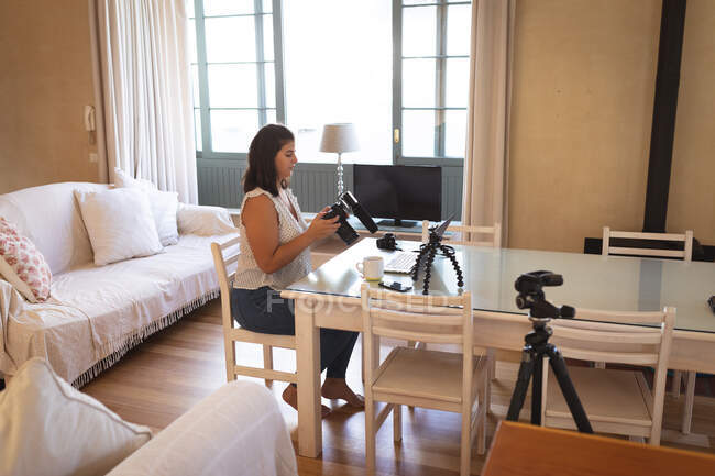 Vlogger mujer caucásica en casa, en su sala de estar con una cámara y un ordenador portátil para preparar su blog en línea. Distanciamiento social y autoaislamiento en cuarentena. - foto de stock