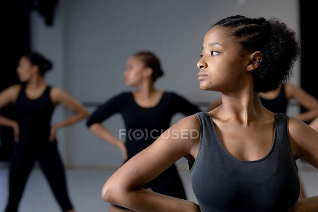 Vista frontal de una bailarina moderna de raza mixta vestida con ropa negra, de pie frente a un grupo multiétnico de bailarinas en forma, sosteniendo sus manos sobre sus caderas. - foto de stock