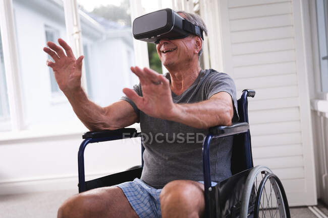 Ein älterer kaukasischer Rentner sitzt an einem sonnigen Tag im Rollstuhl in Unterwäsche vor einem Fenster und benutzt ein VR-Headset mit ausgestreckten Armen vor sich, um sich während der Coronavirus-Pandemie selbst zu isolieren 19 — Stockfoto
