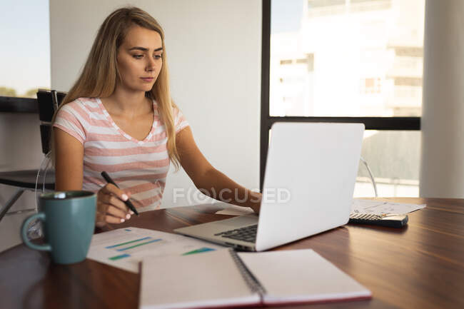 Mujer caucásica sentada junto a una mesa, usando un portátil y escribiendo en una hoja de papel. Distanciamiento social y autoaislamiento en cuarentena. - foto de stock