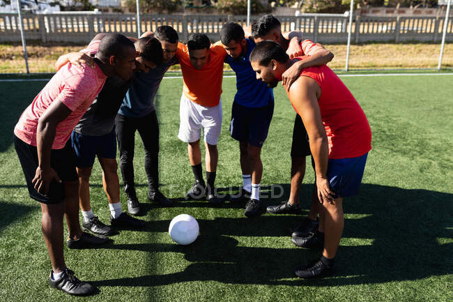 Multi grupo étnico de cinco hombres un lado jugadores de fútbol con entrenamiento de ropa deportiva en un campo de deportes en el sol, de pie en el grupo de motivación antes de una pelota de juego en el medio. - foto de stock