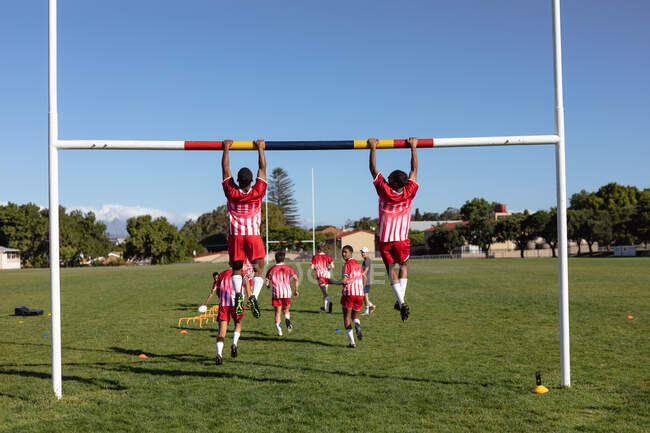 Rear view of two teenage mixed race male rugby players wearing red and white team strip, doing pull-ups hanging from the goal on a rugby pitch, with other players running in the background — Stock Photo