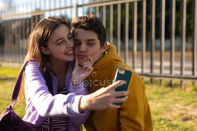 На переднем плане кавказская девочка-подросток и мальчик обнимаются и улыбаются, девушка сжимает лицо мальчика, пока она делает селфи с их смартфоном на территории школы, другая подростковая пара сидит на заднем плане — стоковое фото
