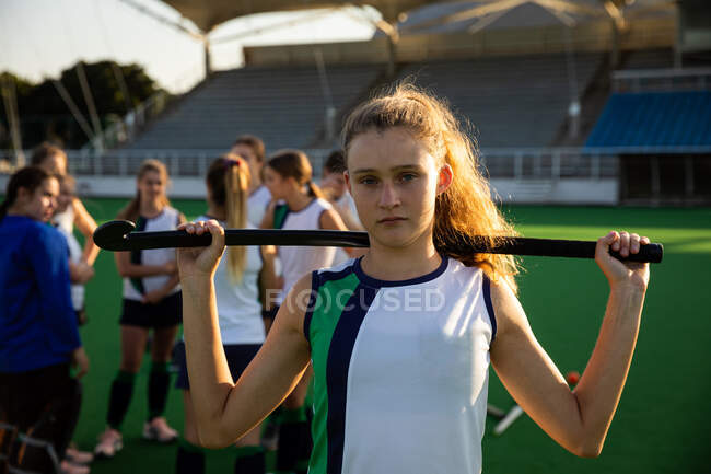 Portrait of a Caucasian female field hockey player, preparing before a game, standing on a hockey pitch, looking at camera, holding a hockey stick on her shoulders, with her teammates standing in the background on a sunny day — Stock Photo