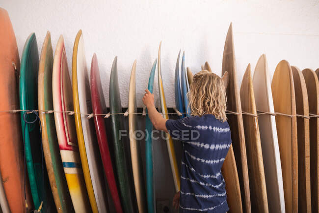 Задний вид кавказского серфингиста в его студии, проверяющего одну из досок для сёрфинга стоящей, с досками для сёрфинга в стойке позади него. — стоковое фото
