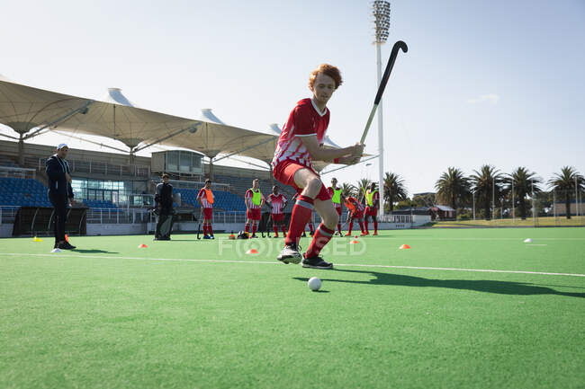 Side view of a teenage Caucasian male field hockey player, training on the hockey pitch before a game, preparing to hit the ball with a hockey stick, with his teammates and coach in the background on a sunny day — Stock Photo