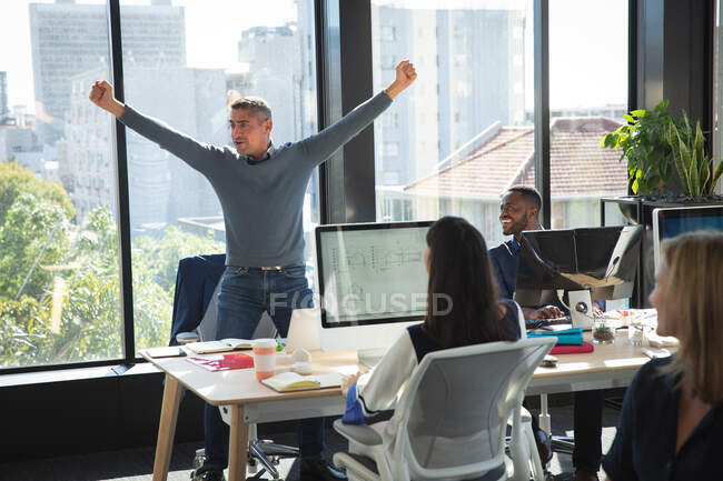 Кавказский бизнесмен, работающий в современном офисе, стоя и празднуя, держа руки в воздухе, со своими коллегами, работающими на заднем плане — стоковое фото