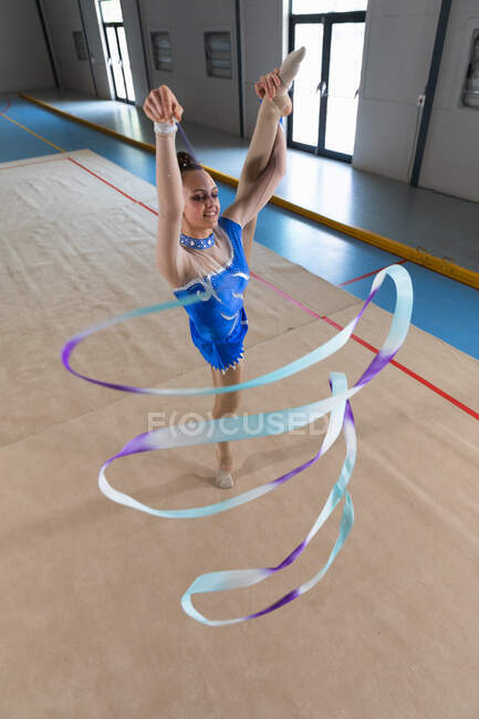 Vista frontal de ángulo alto de la gimnasta femenina caucásica adolescente que realiza en el gimnasio, haciendo ejercicio con cinta, de pie en split, un brazo extendido, usando maillot azul - foto de stock