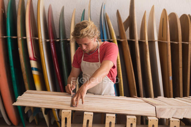 Кавказький самець серфінгу працює в своїй майстерні, вирізає дерев'яні смуги і готується зробити серфінг, з серфінгом на стійці на задньому плані. — стокове фото