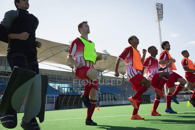 Side view of a multi-ethnic group of teenage male field hockey players training before a game, working out on a field hockey pitch, running on the spot on a sunny day — Stock Photo