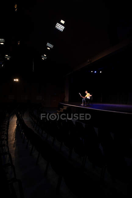 Вигляд на Кавказького підлітка-студента старшокласника в порожньому шкільному театрі, який готується перед виступом, сидить на стільці на сцені, тримає сценарій і практикує свою роль — стокове фото